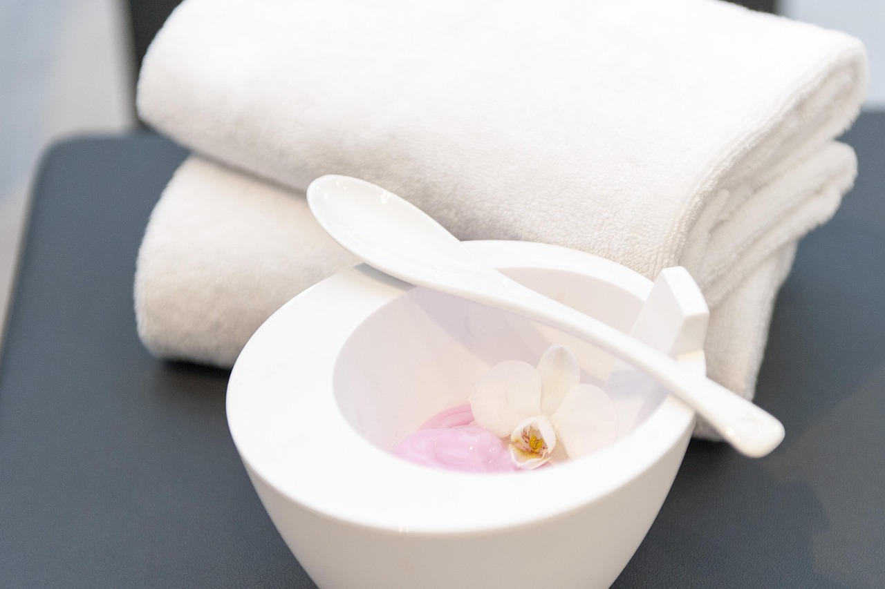 A white bowl and spatula for mixing a face mask beside rolled towels